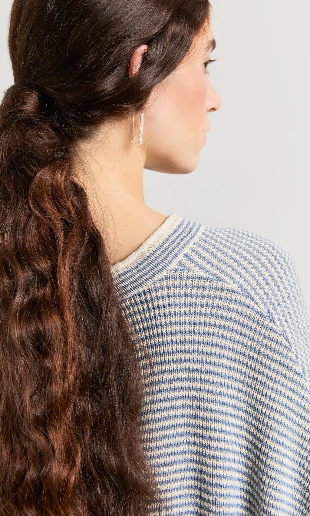 Plümo- Woman with long wavy brown hair in a loose ponytail wears the Boathouse Jumper, seen from behind.