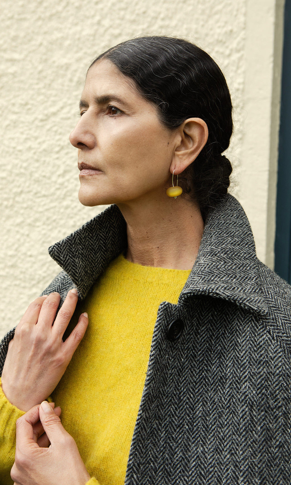 Plümo  Woman in a yellow sweater and gray coat, wearing Bola Earrings - Honey Dew, stands by a textured wall.