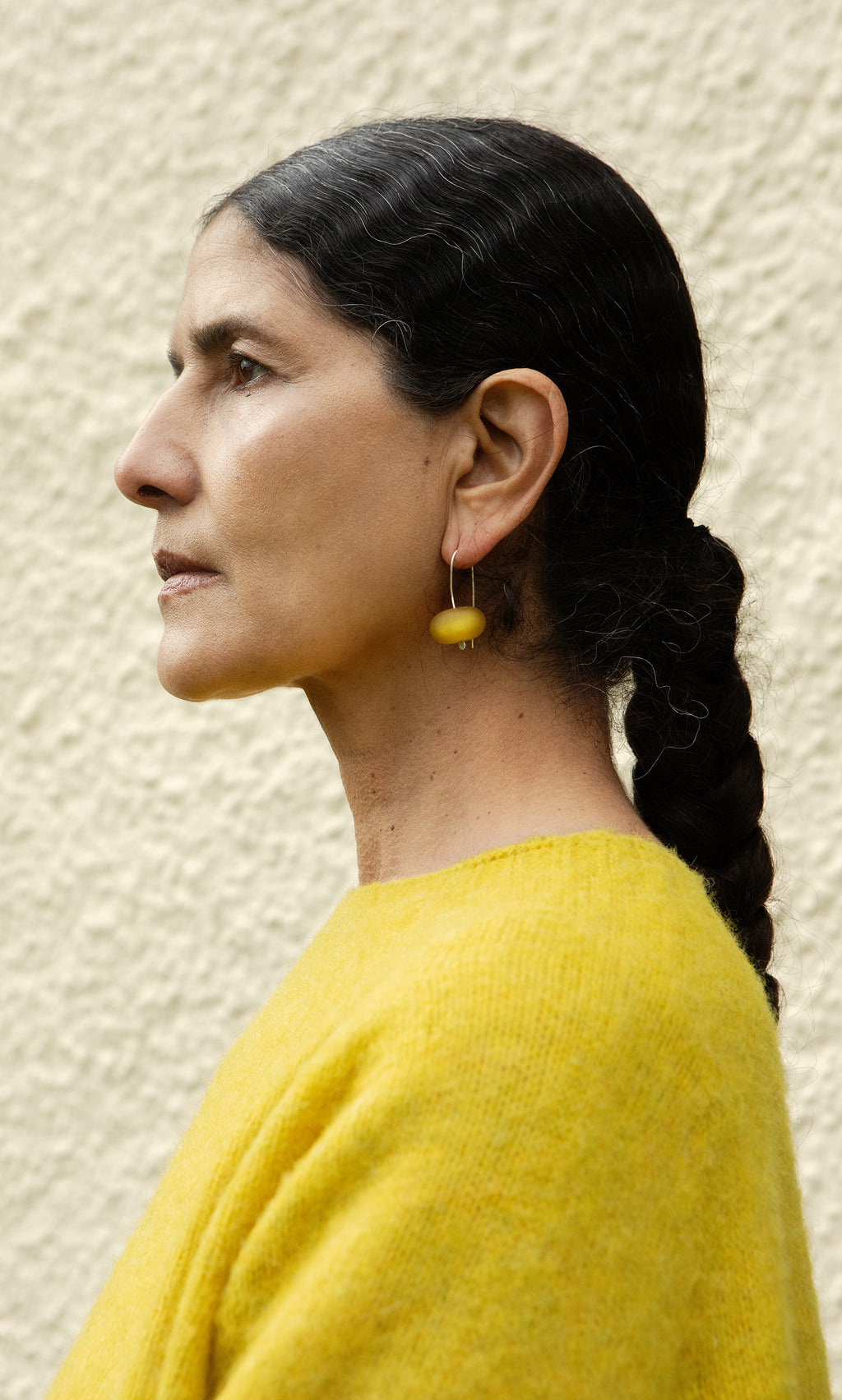 Plümo  Woman in profile with dark braids in a yellow top and Bola Earrings - Honey Dew, against a textured wall.