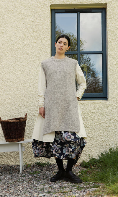 Plümo  A person in layers stands by a window, wearing a gray vest, cream dress, and the Pugh Flower Skirt.