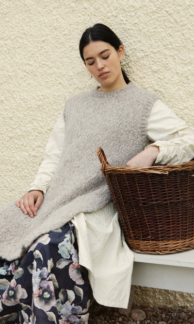 Plümo  A woman in a gray Borwick Tunic and floral skirt sits on a bench, holding a wicker basket with her eyes closed.