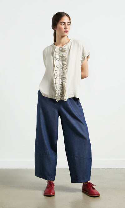 Plümo- A woman in a relaxed fit wears the Birch Ruffle Top with blue pants and red shoes against a plain white wall.