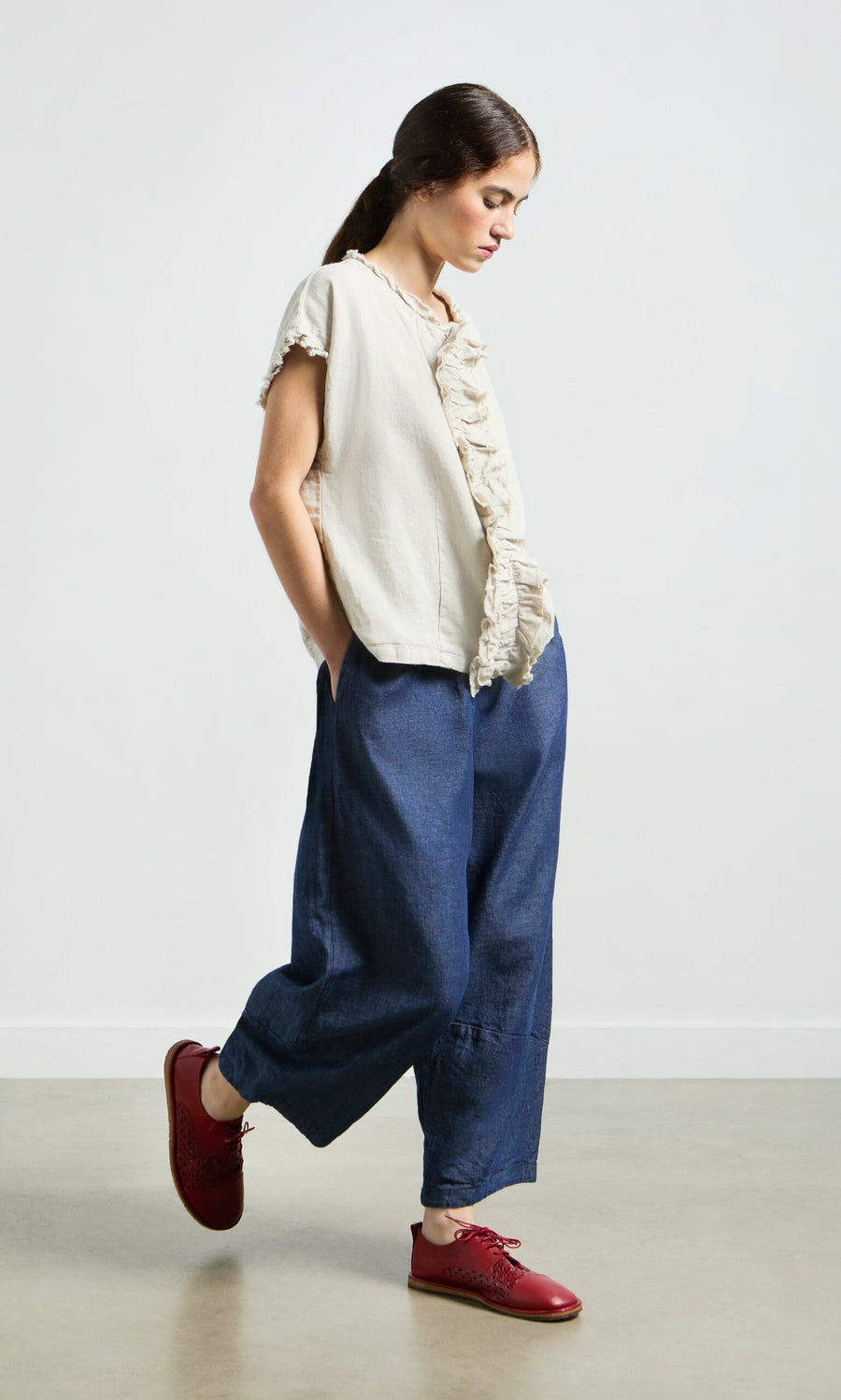 Plümo- Woman in a cream ruffled blouse and Mara Barrel Pants stands with hands in pockets on a plain background.