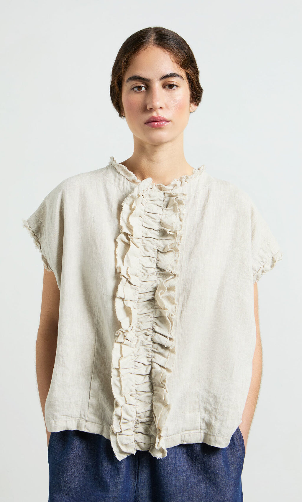 Plümo- Woman wearing the Birch Ruffle Top and blue pants, standing against a plain light background.