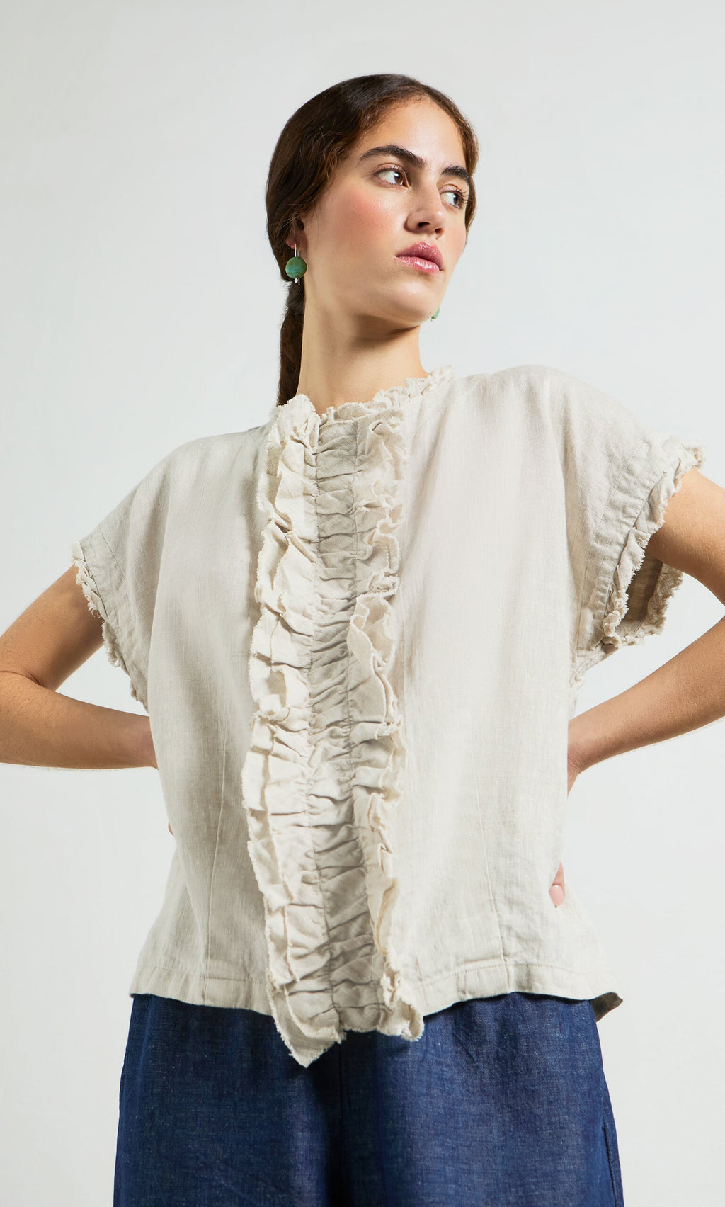Plümo- Woman in a relaxed silhouette Birch Ruffle Top and blue pants stands hands on hips, looking to the side.