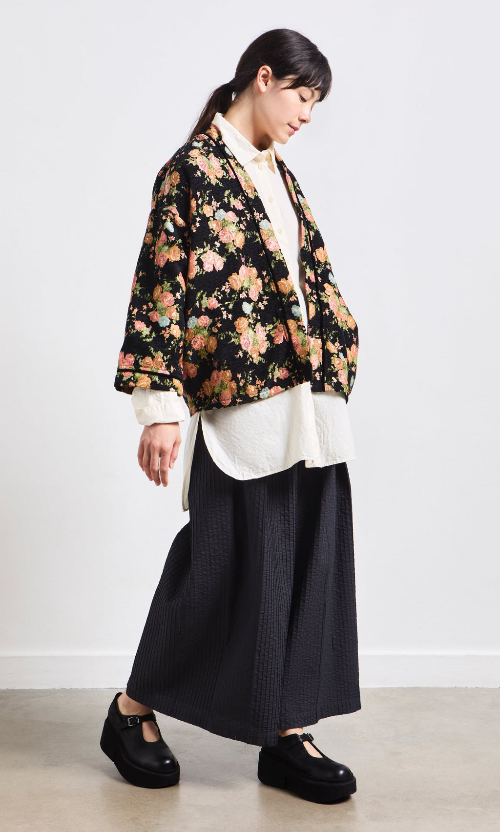 Plümo- Person wearing a floral kimono jacket, white shirt, Kiri Pintuck Pants, and black shoes walking indoors.