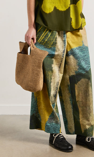 Plümo- Person wearing Brushstroke Pants with an elasticated drawstring waist, black shoes, tan woven bag, and green top.