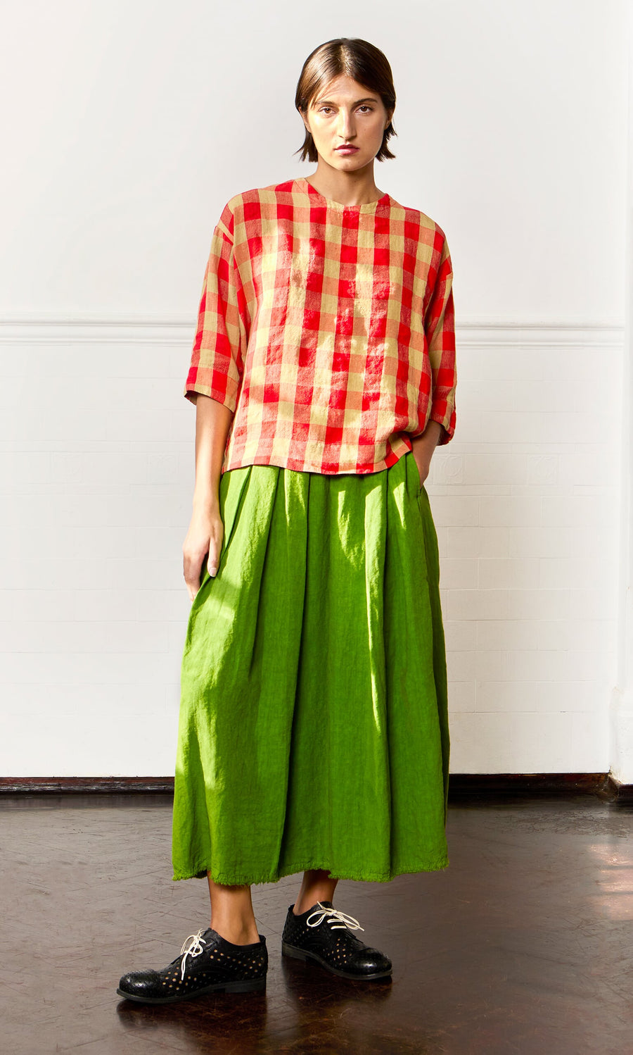 Plümo- Person wears the Burgos Blouse in red and cream gingham, a bright green skirt, and black shoes against a white wall.