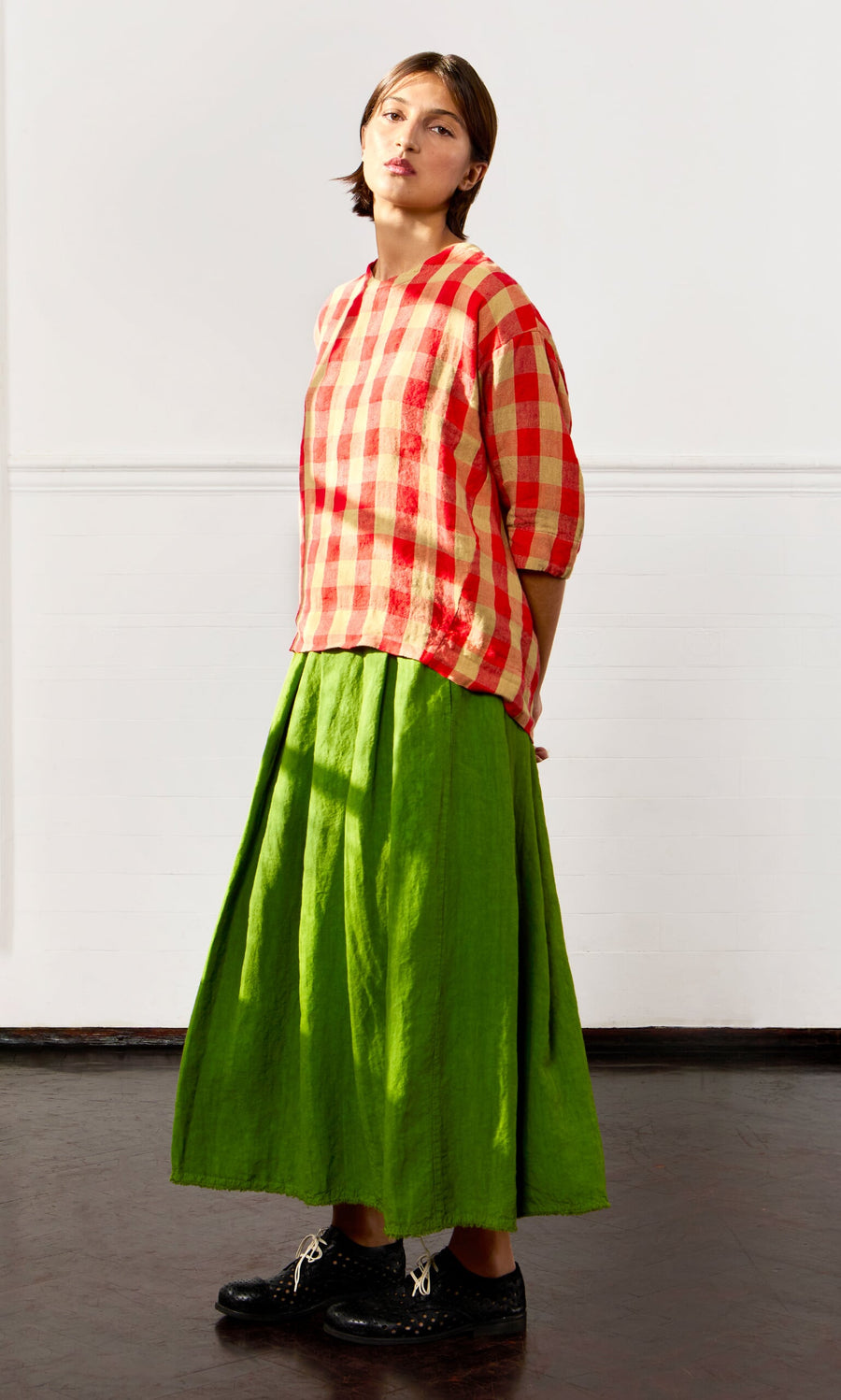 Plümo- Woman in a Burgos Blouse, green long skirt, and black shoes stands against a plain white indoor wall.