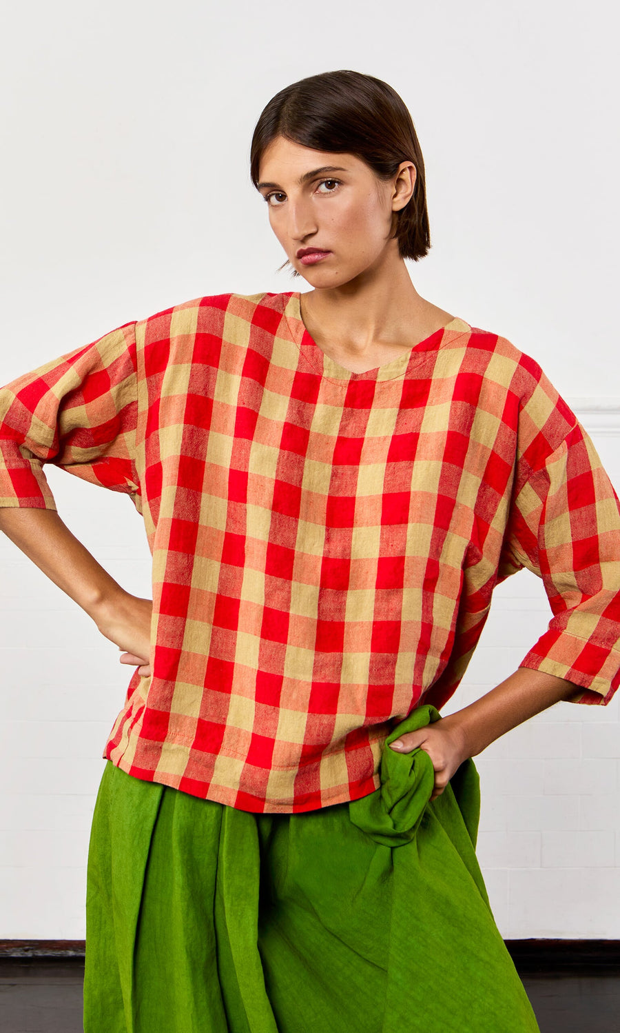 Plümo- Person wearing the Burgos Blouse in red and beige gingham with bright green pants, hand on hip against a plain background.