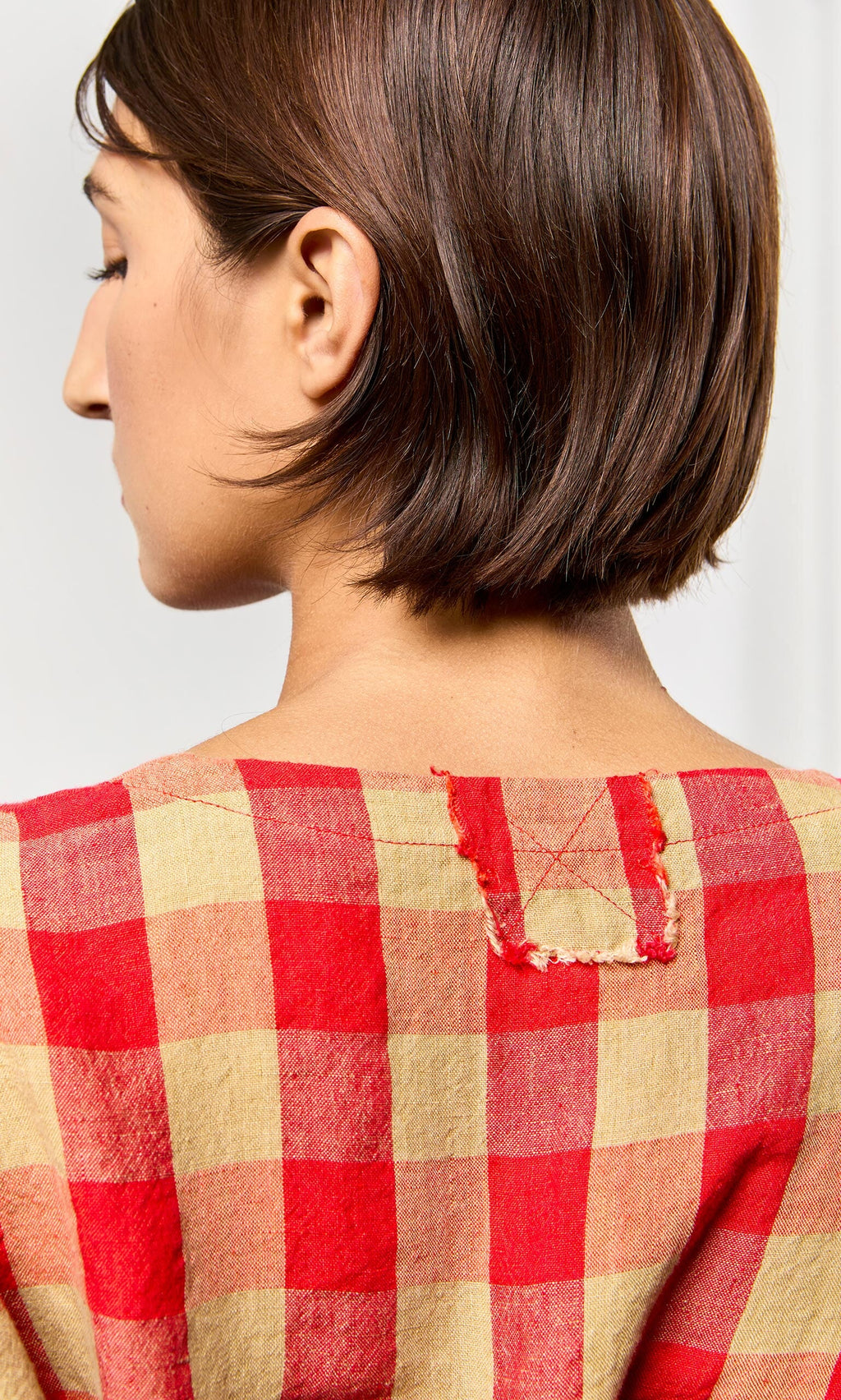 Plümo- Woman with short brown hair wears the Burgos Blouse in gingham check, shown from the back with a visible fabric patch.
