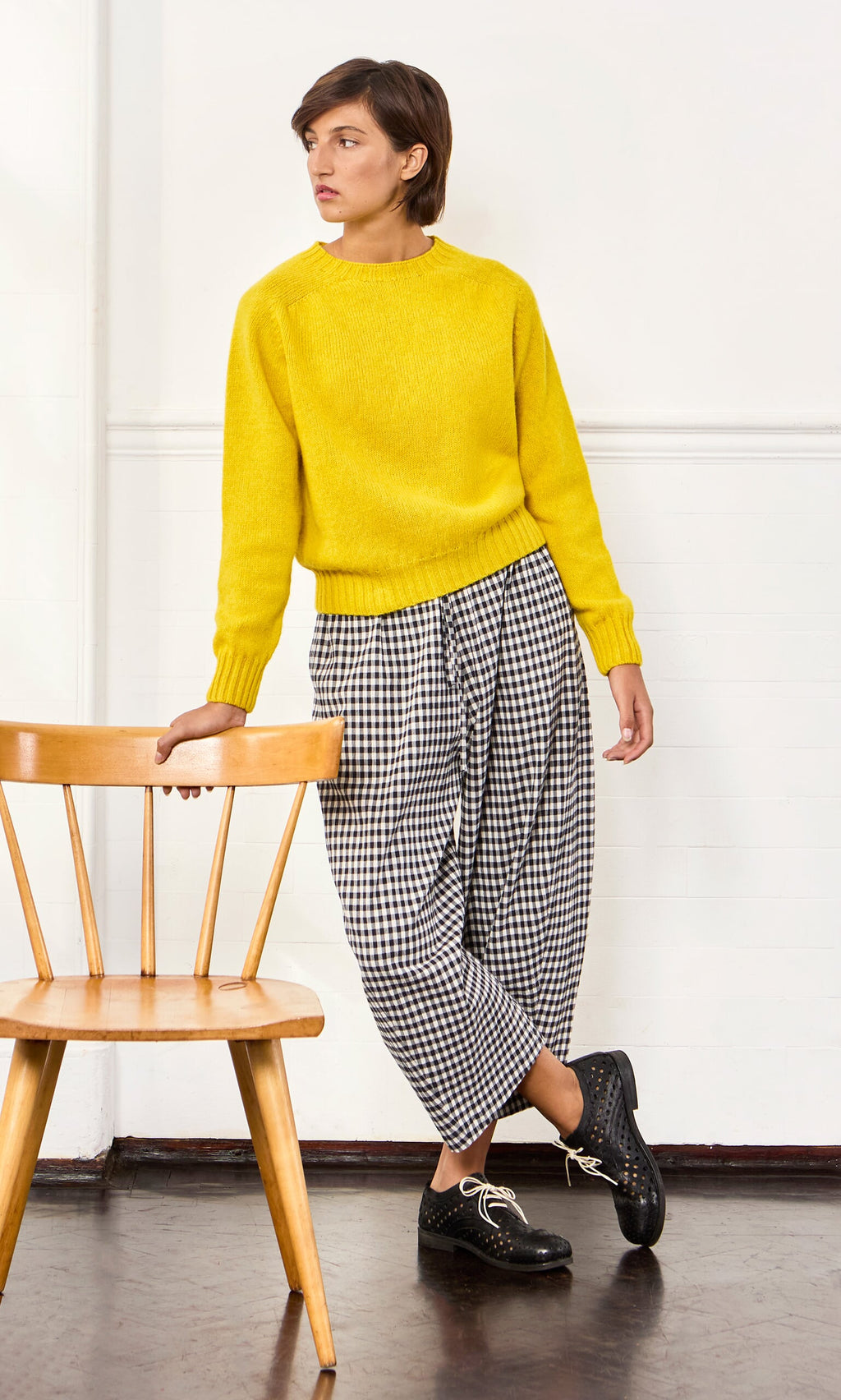 Plümo- Person in Butoni Pants - Black and White leans on a wooden chair, looking to the side in a minimal room.