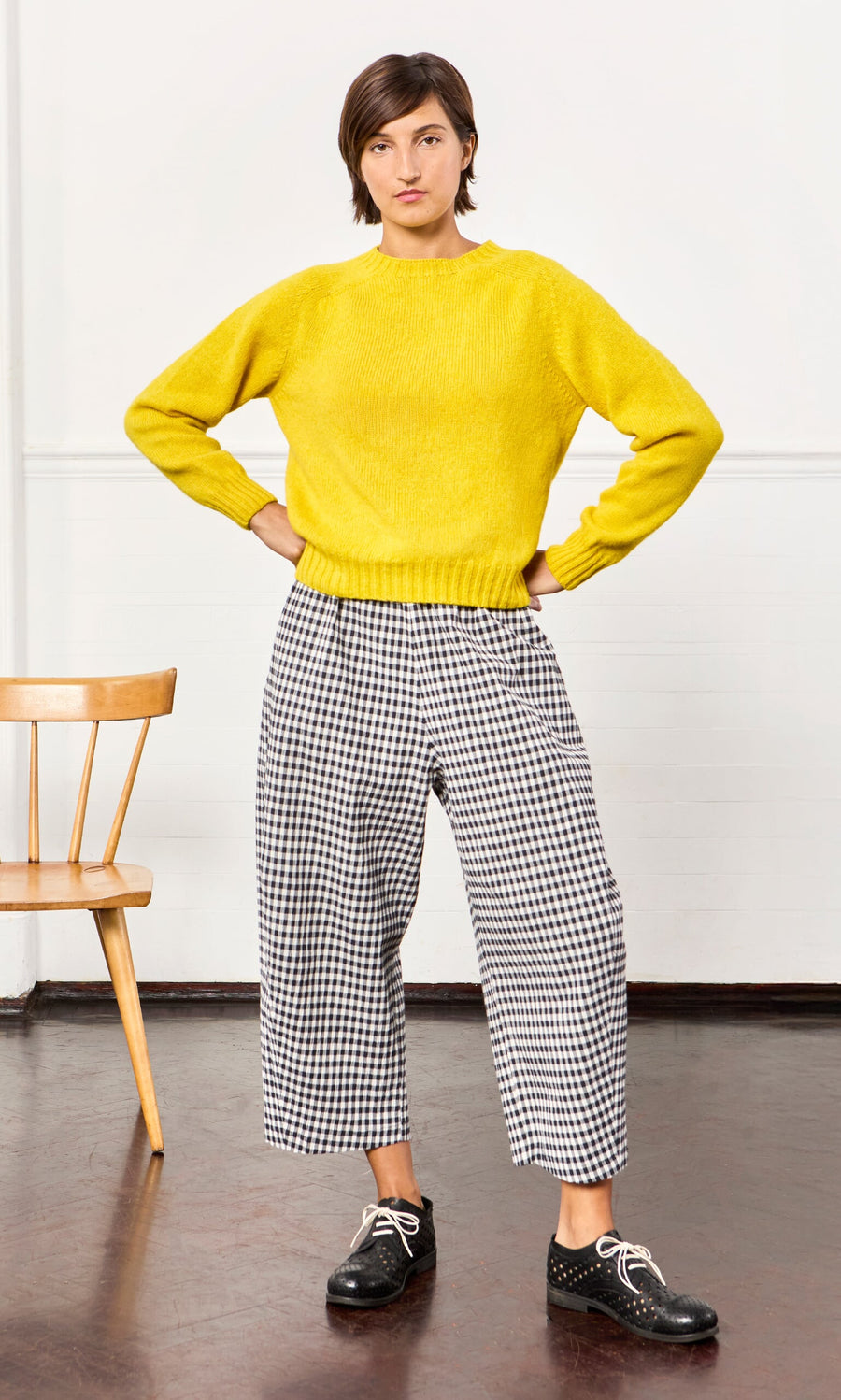 Plümo- Person indoors in a yellow sweater, Butoni Pants - Black and White with drawstring waist, black shoes, wooden chair nearby.
