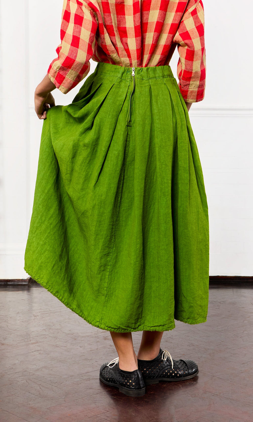 Plümo- Person in a red plaid shirt, Cadiz Skirt in bright green linen, and black shoes stands on a dark floor.