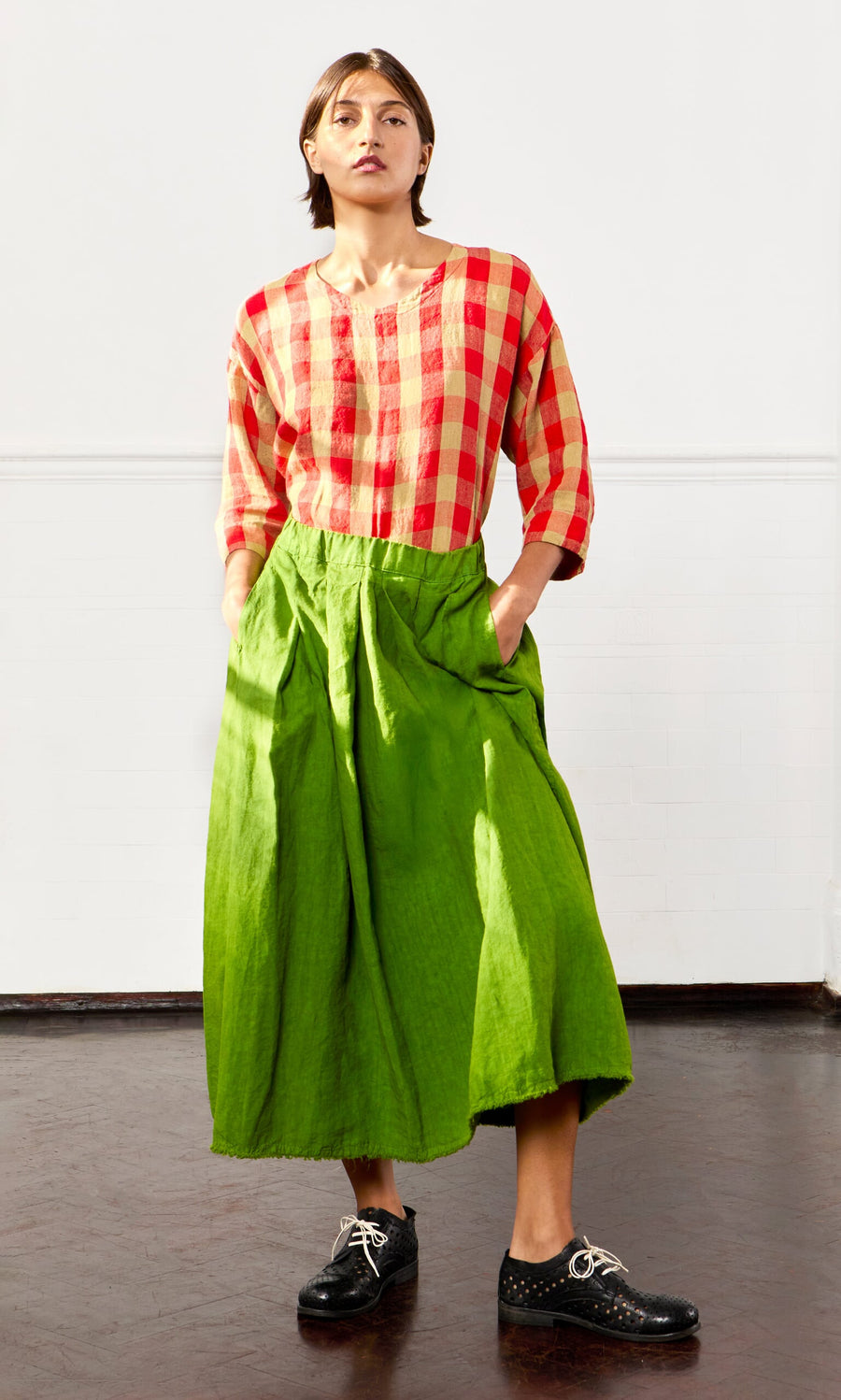 Plümo- A person stands with hands in pockets, wearing the Cadiz Skirt, a red checkered shirt, and black shoes.