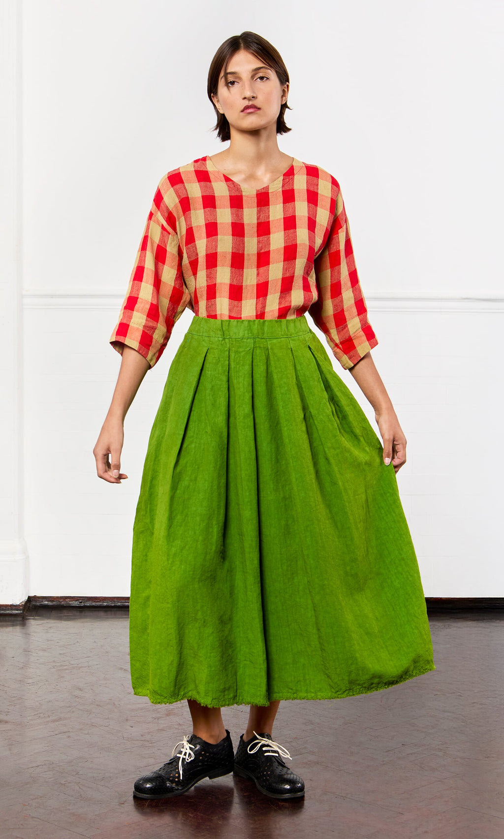 Plümo- Woman in a red checkered top, Cadiz Skirt, and black shoes stands against a plain white wall.