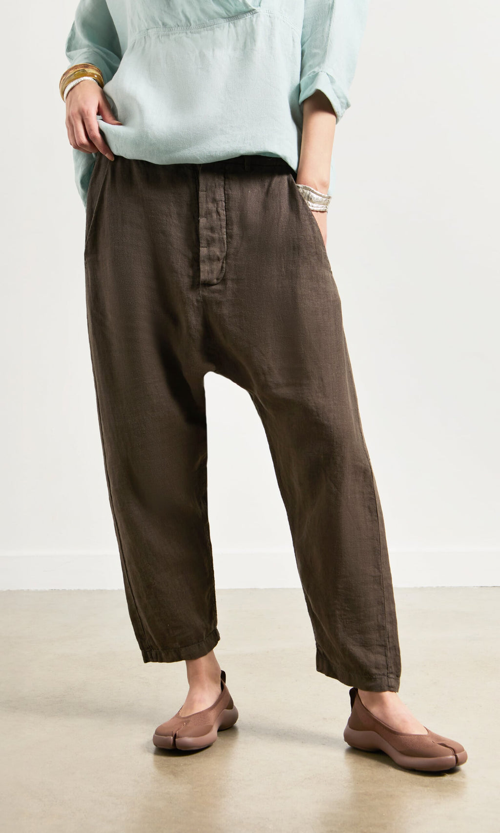Plümo- Person wears Caper Pants - Brown, a relaxed linen chino, with a light blue top, beige shoes, and bracelets.
