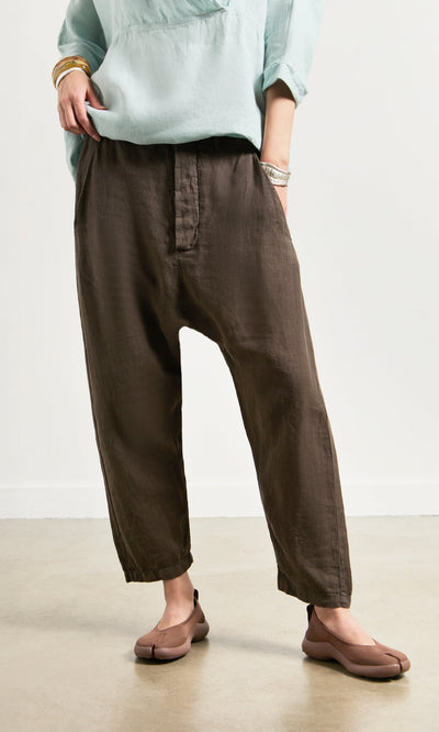 Plümo- Person wears Caper Pants - Brown, a relaxed linen chino, with a light blue top, beige shoes, and bracelets.