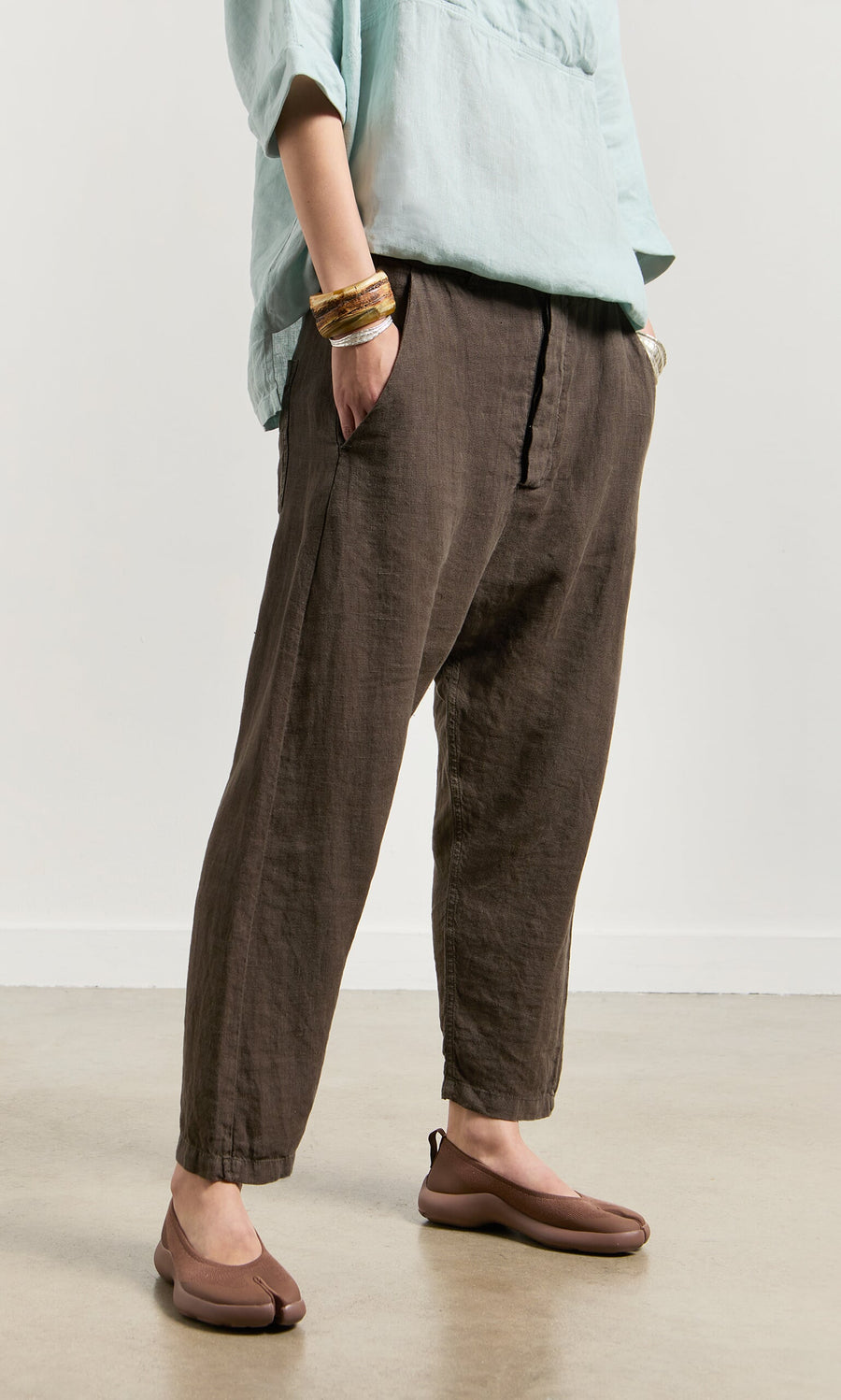 Plümo- Person wears Caper Pants - Brown, light blue top, brown shoes, and bracelets, standing hands in pockets.