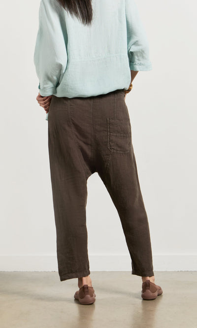 Plümo- Person stands with back to camera in a light blue top, Caper Pants - Brown, and brown sandals on a light floor.