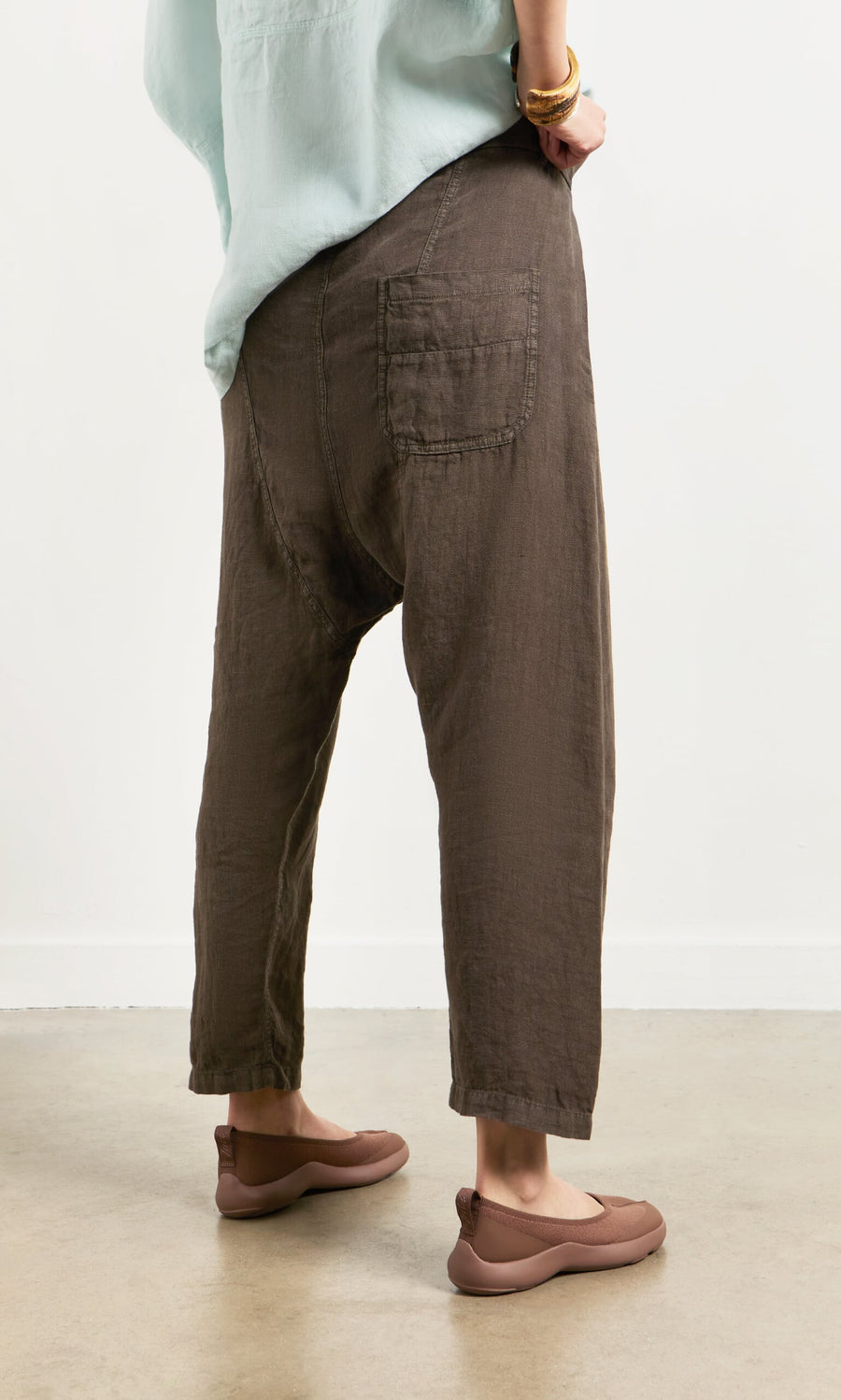 Plümo- Person wearing Caper Pants - Brown with taupe flats, seen from behind, standing on a grey floor.