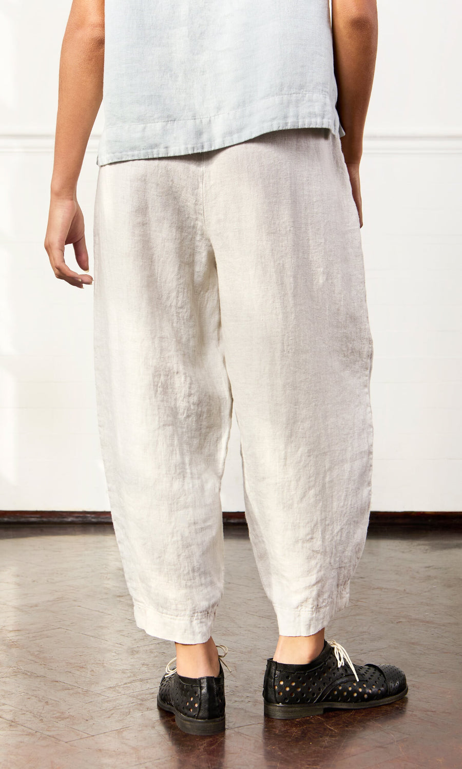 Plümo- Person indoors wearing Cazalla Pants—loose, sustainably made white linen with elastic waist—and a light blue top.