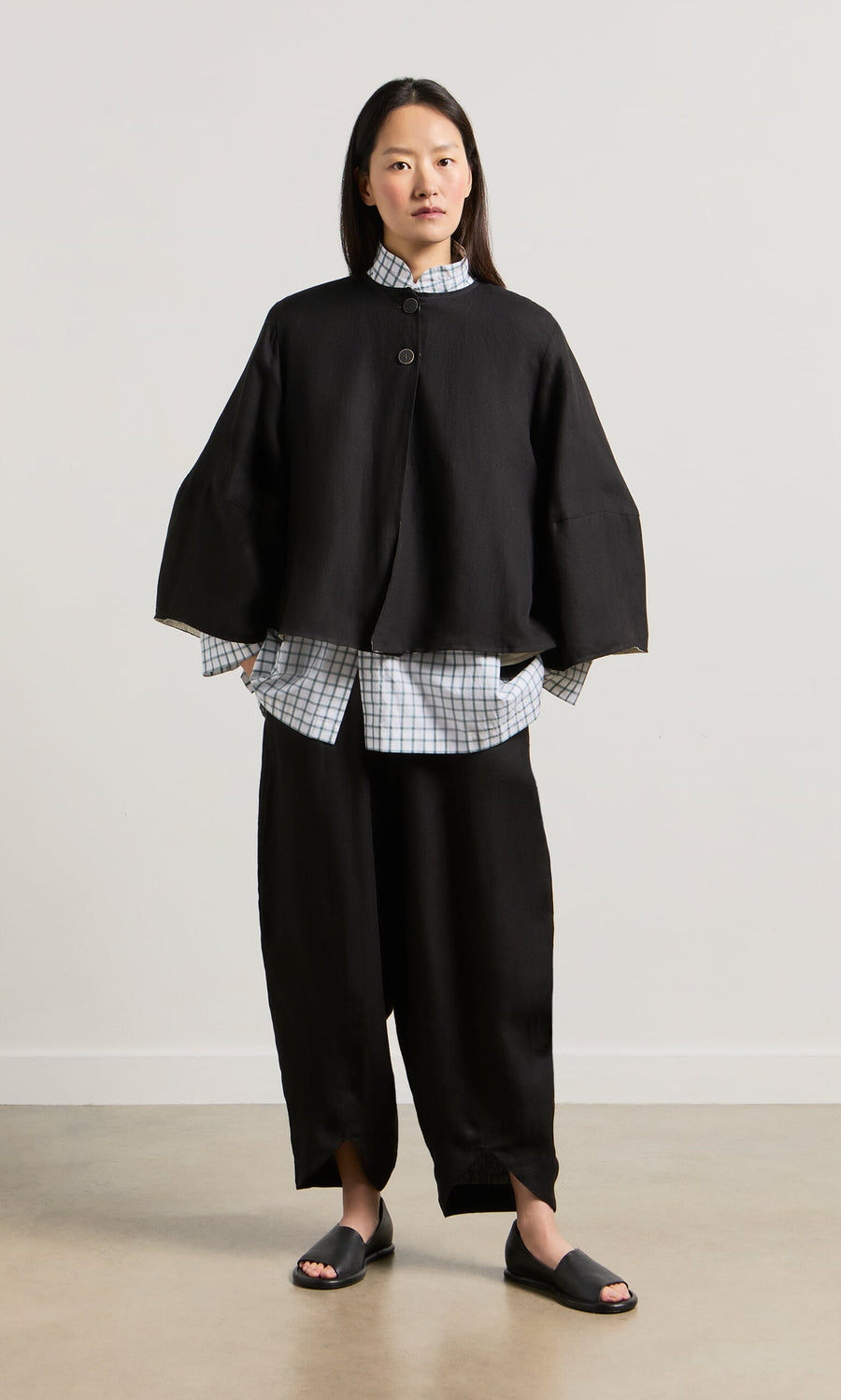 Plümo- Woman wearing the Holden Jacket with bell sleeves over a checked shirt, black loose pants, and slide sandals.