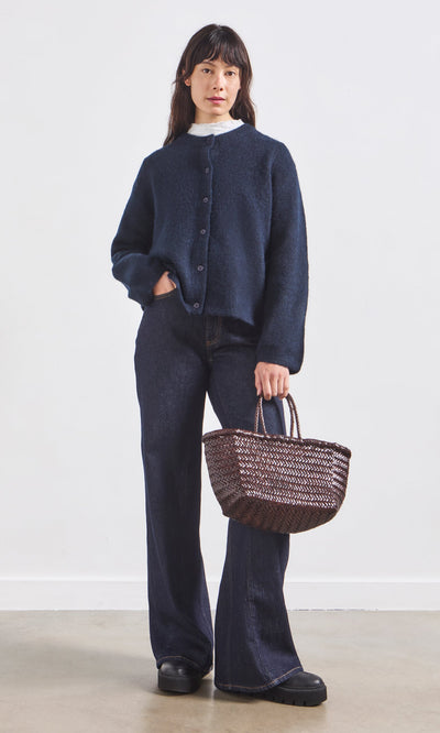 Plümo- A woman in Belted Wide Jeans, a navy cardigan, and black shoes holds a woven basket bag against a plain background.
