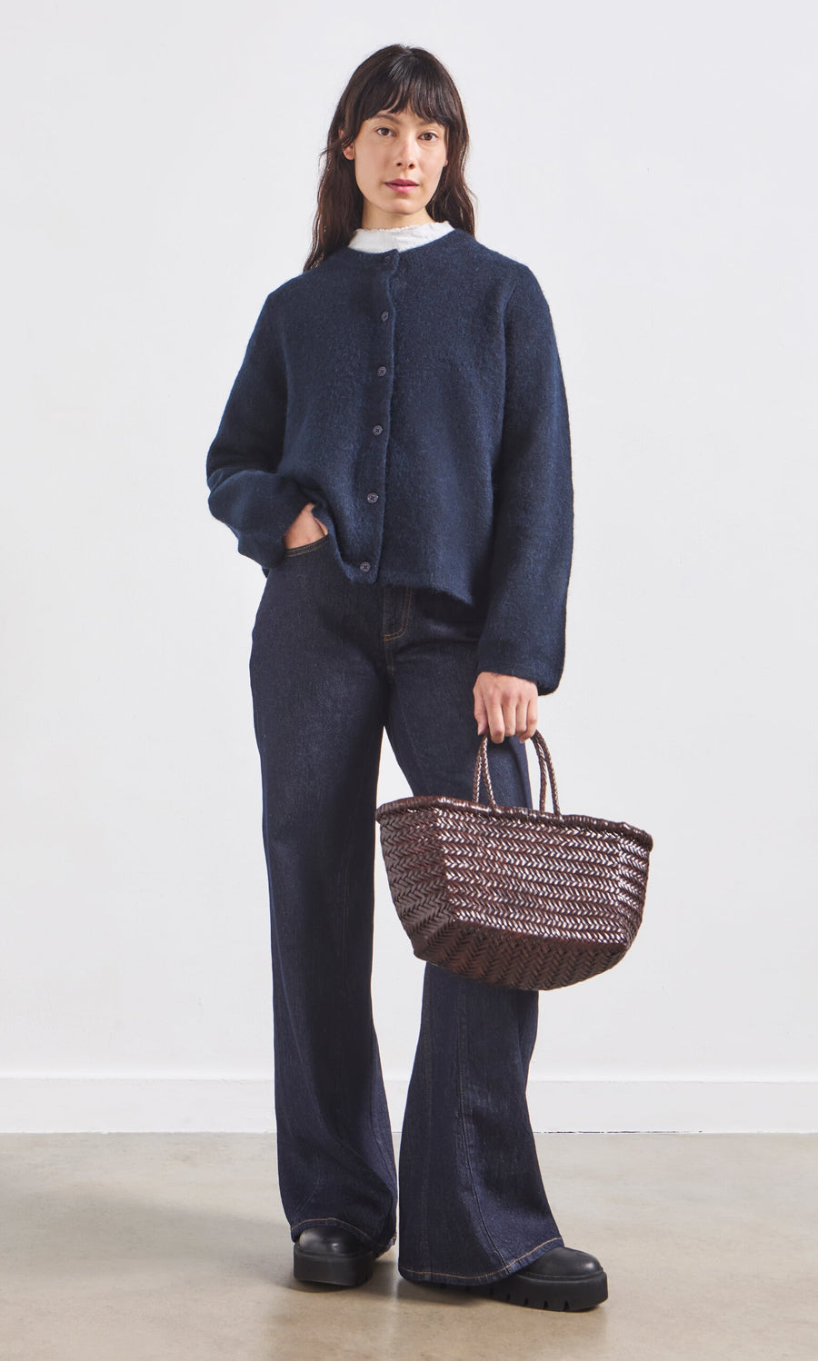 Plümo- A woman in Belted Wide Jeans, a navy cardigan, and black shoes holds a woven basket bag against a plain background.