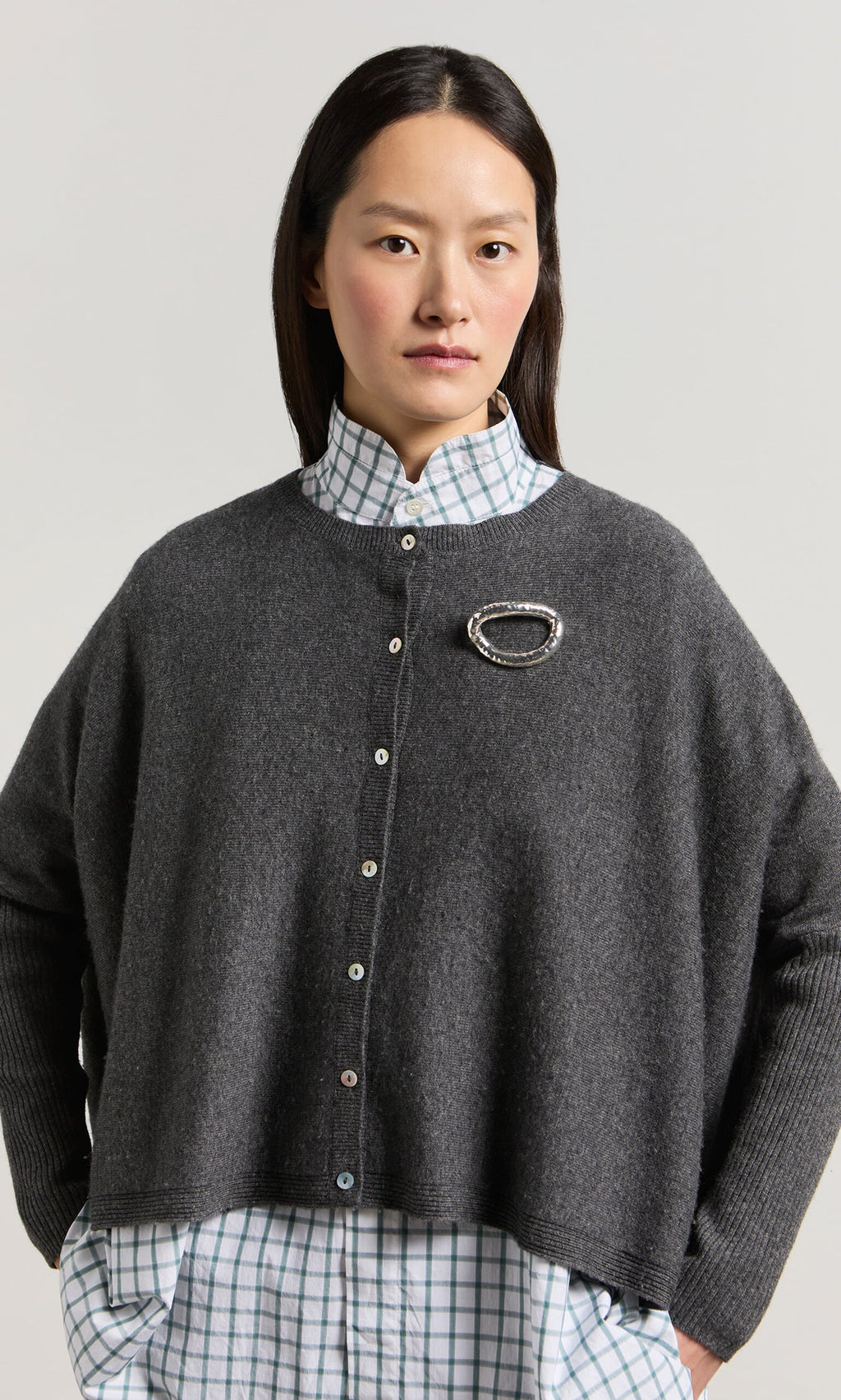 Plümo- Woman wears the Alma Cardigan - Grey with rib-knit sleeves over a white and green plaid shirt, brooch attached.