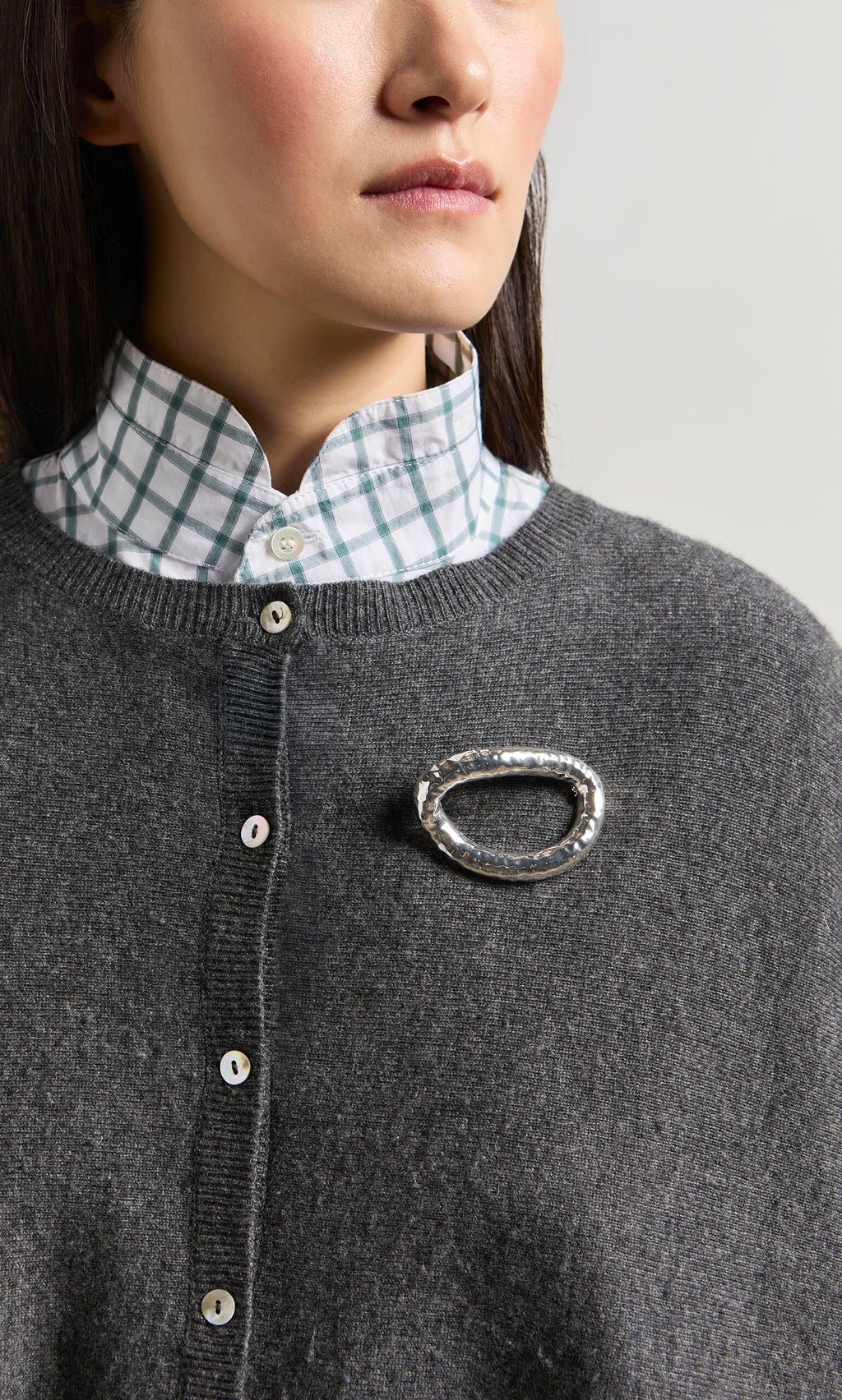 Plümo- Woman wearing a grey sweater, plaid shirt, and the Circle Brooch, an elliptical silver accessory with a textured finish.