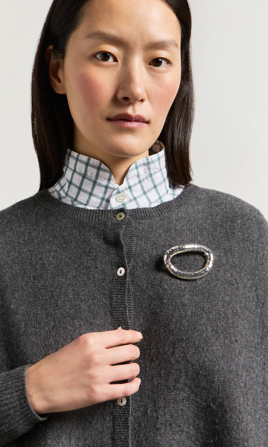 Plümo- Woman wearing a gray cardigan, checkered shirt, and Circle Brooch, looking directly at the camera.