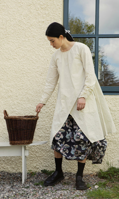 Plümo  Person in a long cream dress and Pugh Flower Skirt stands outside, hand on a wicker basket by a window.
