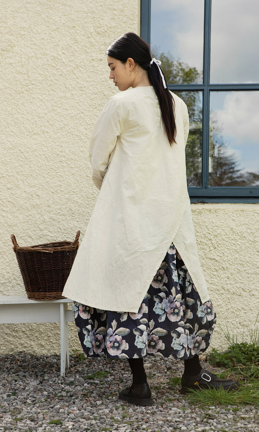 Plümo  Woman in a cream coat, Clementine Blouse, and floral skirt stands by a white bench with a wicker basket.