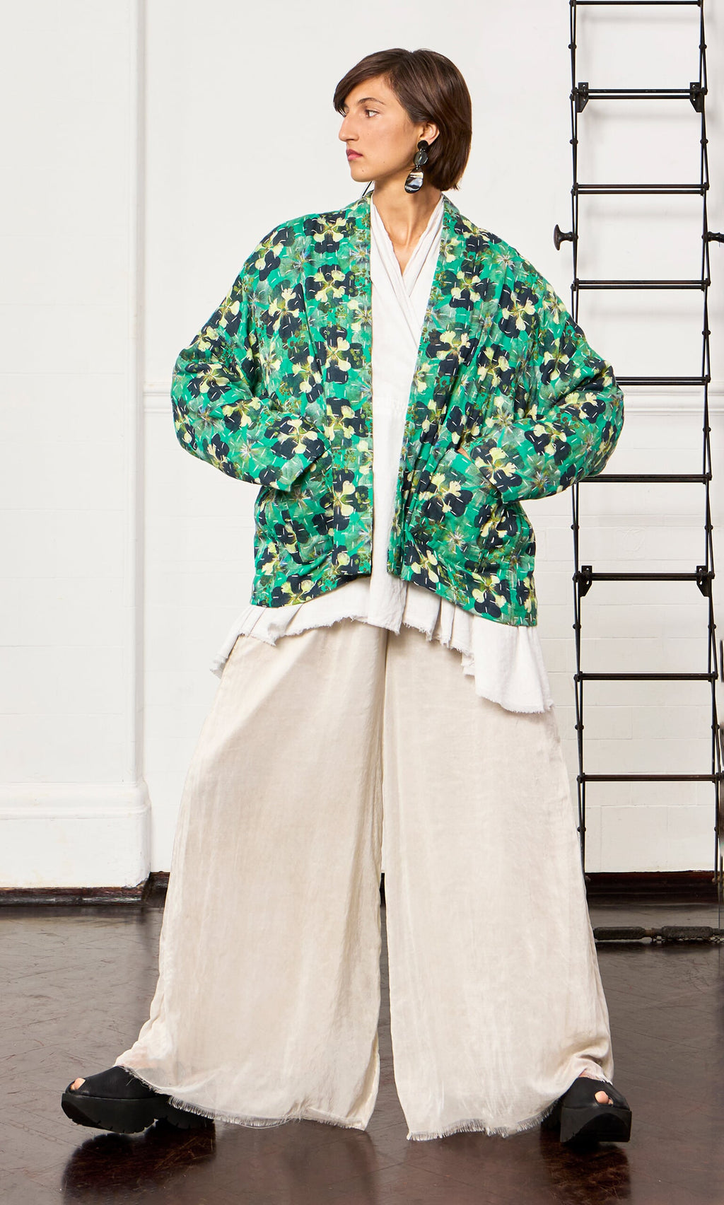 Plümo- Woman in relaxed fit cream pants and the Clover Jacket stands confidently indoors near a black metal ladder.