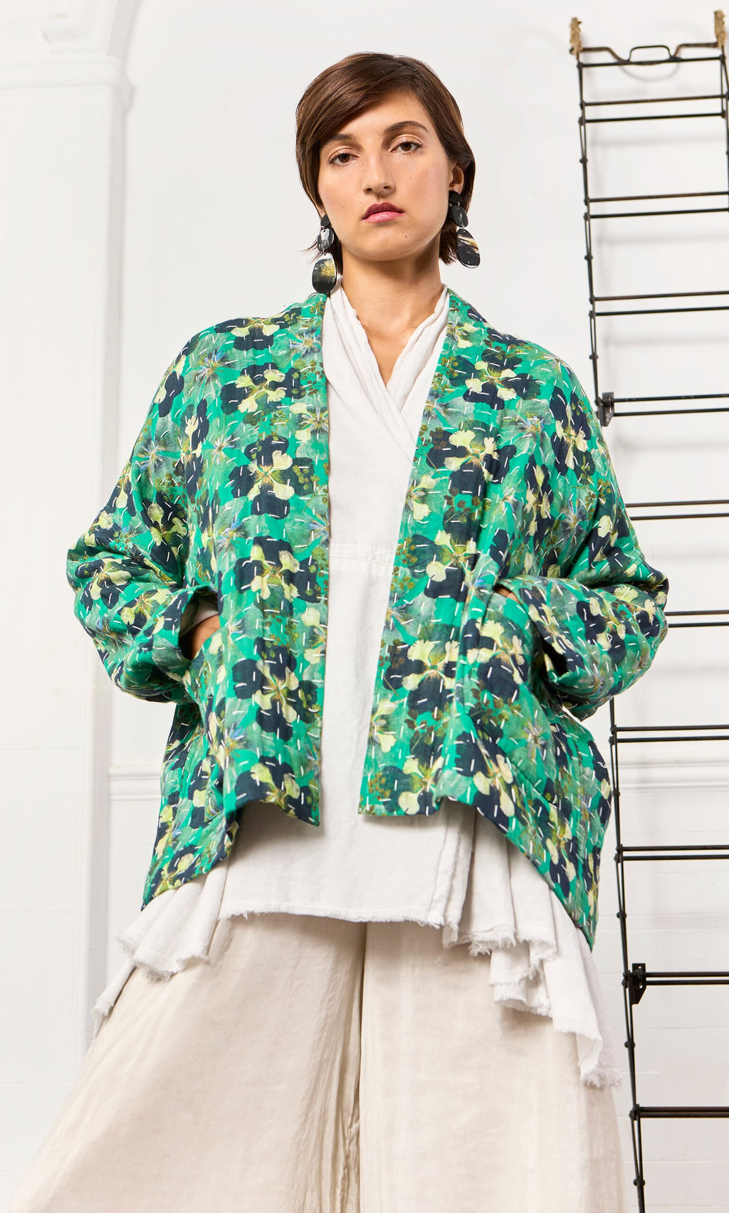 Plümo- Woman models the Clover Jacket, a green pansy print kimono, over white with hands in pockets, exuding confidence.