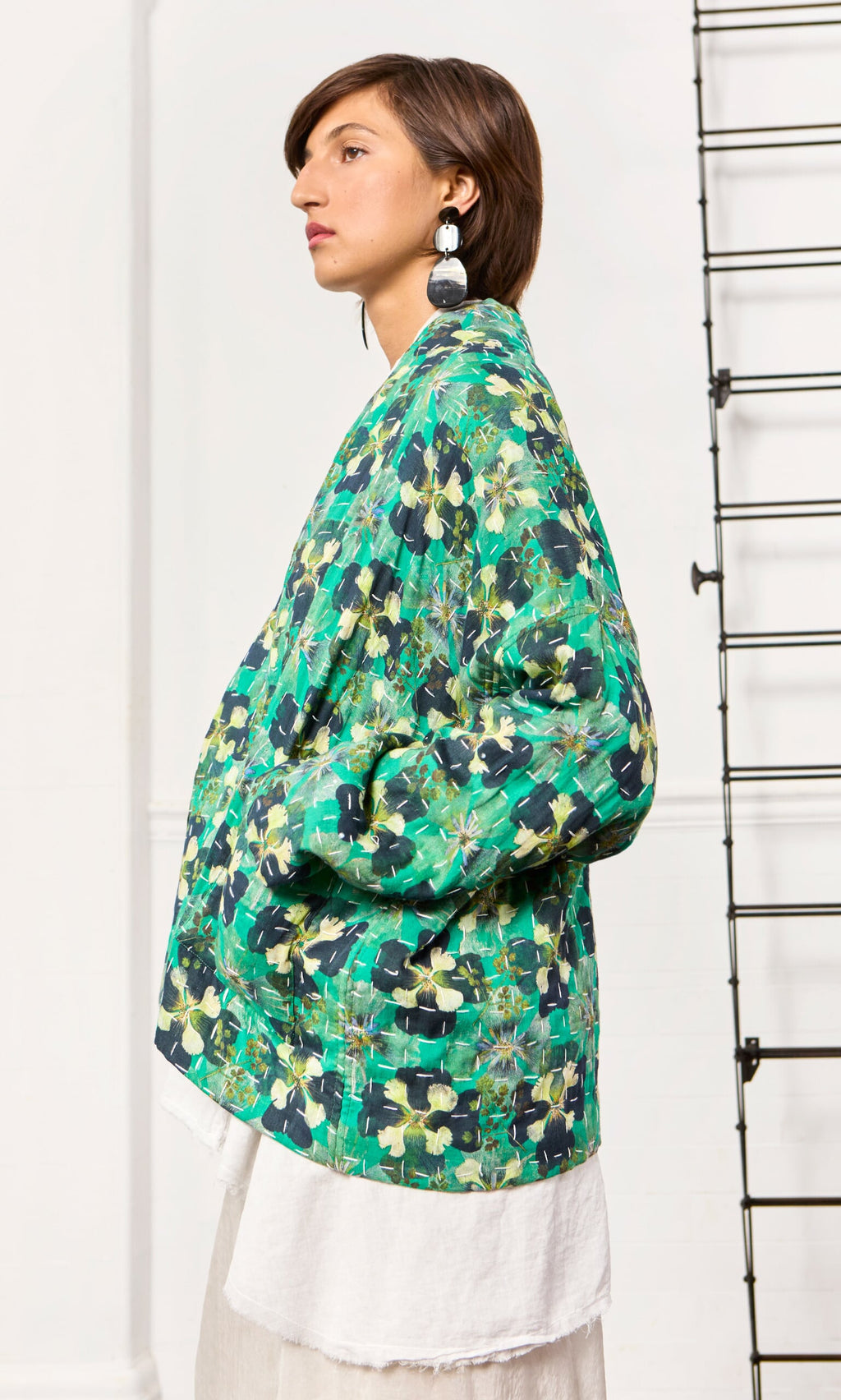 Plümo- Wearing a relaxed fit, green floral Clover Jacket and white skirt, person stands sideways with hands in pockets.