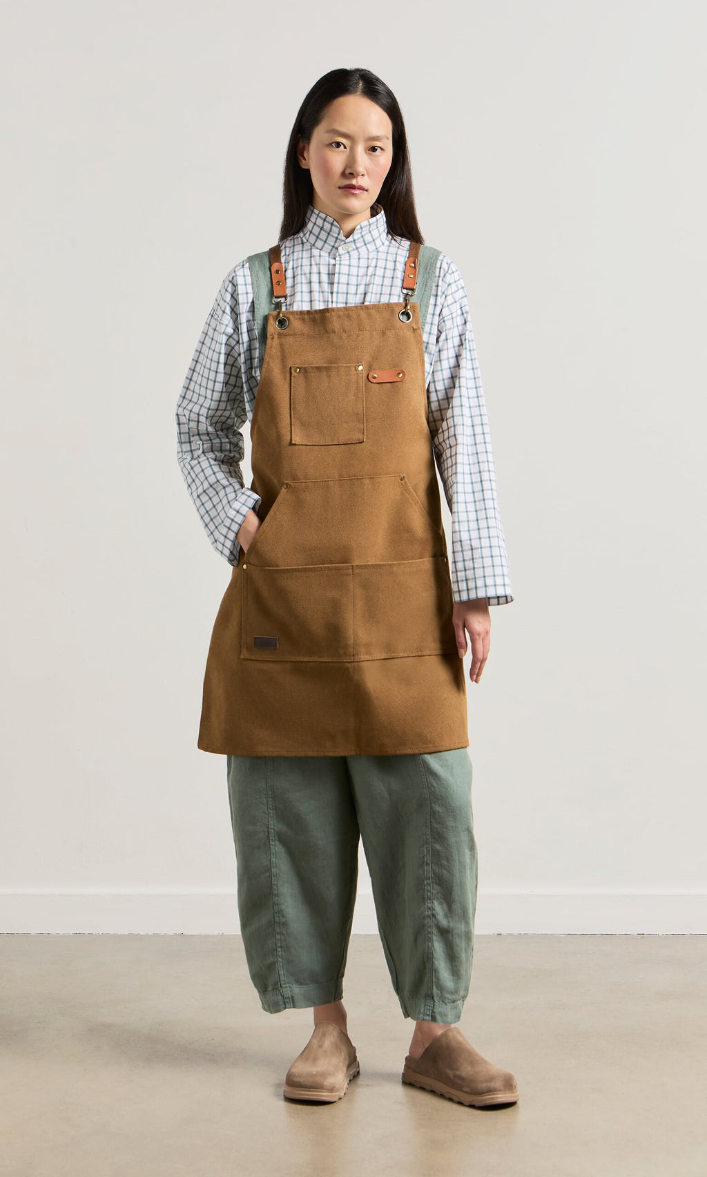 Plümo- Person in a Cobbler Apron with faux leather straps, plaid shirt, green pants, and suede shoes stands against a plain background.
