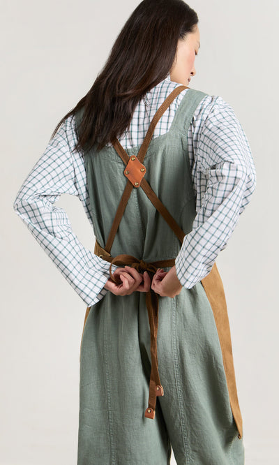Plümo- Woman in a checkered shirt ties faux leather straps on the green and brown Cobbler Apron, facing away.
