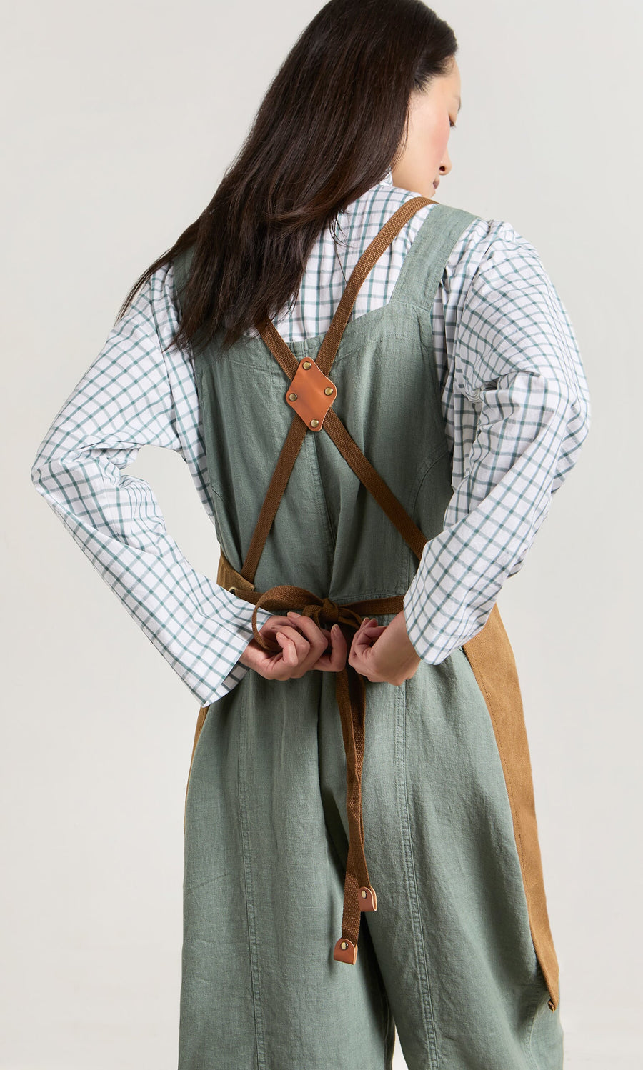 Plümo- Woman in a checkered shirt ties faux leather straps on the green and brown Cobbler Apron, facing away.