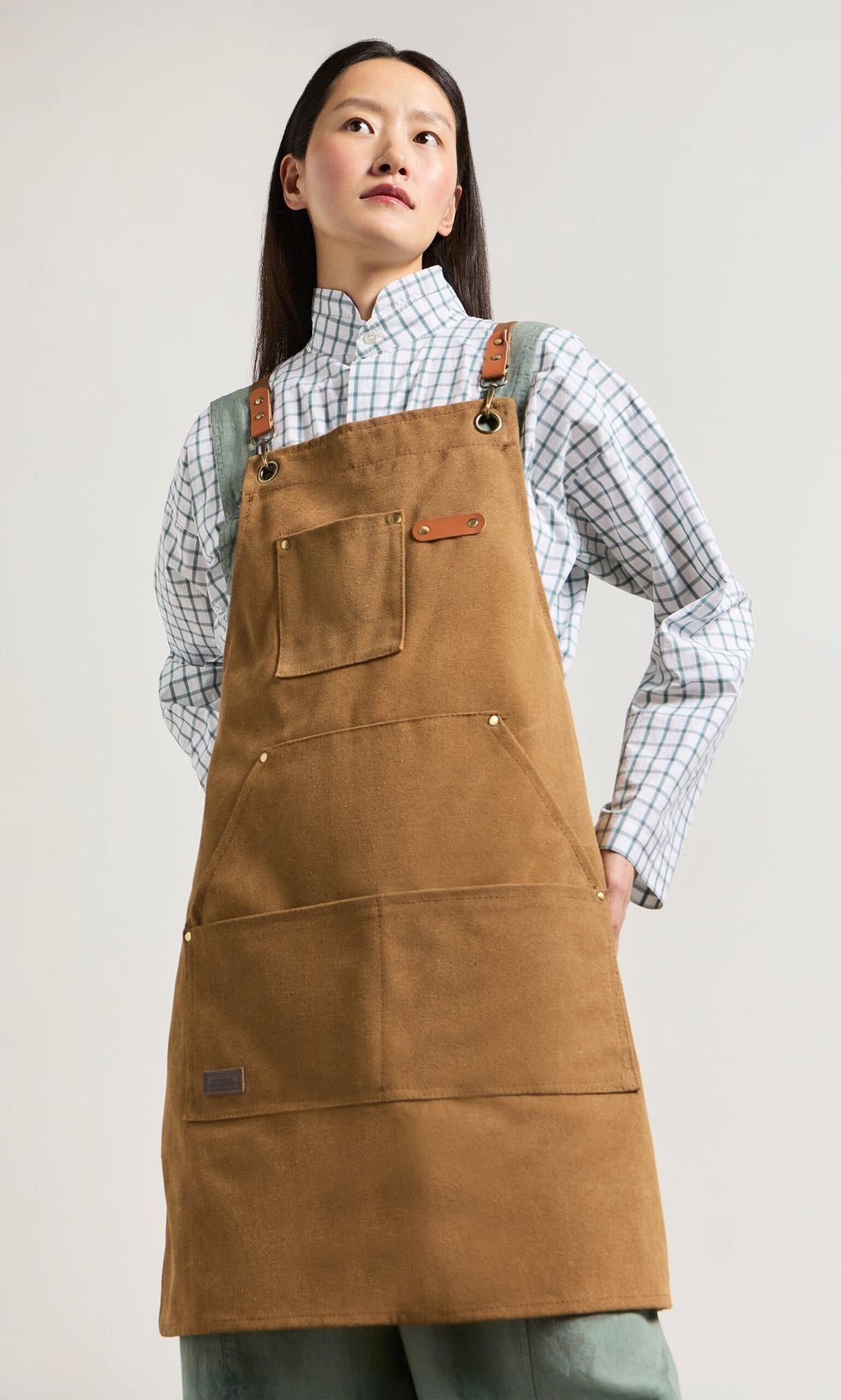 Plümo- Person wears the Cobbler Apron with faux leather straps over a plaid shirt, hands in pockets, looking upward.