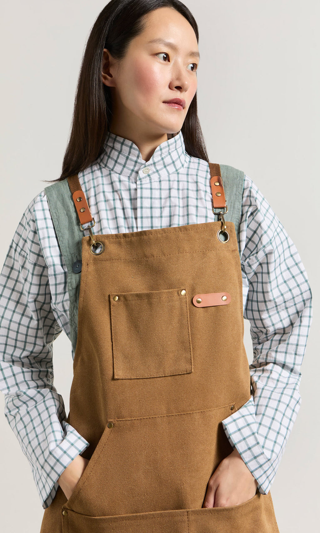Plümo- Person wearing a Cobbler Apron with faux leather straps over a white and green plaid shirt, looking to the side.