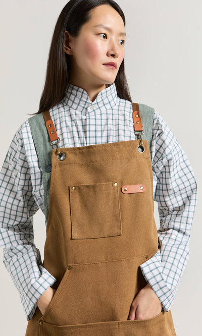 Plümo- Person wearing a Cobbler Apron with faux leather straps over a white and green plaid shirt, looking to the side.
