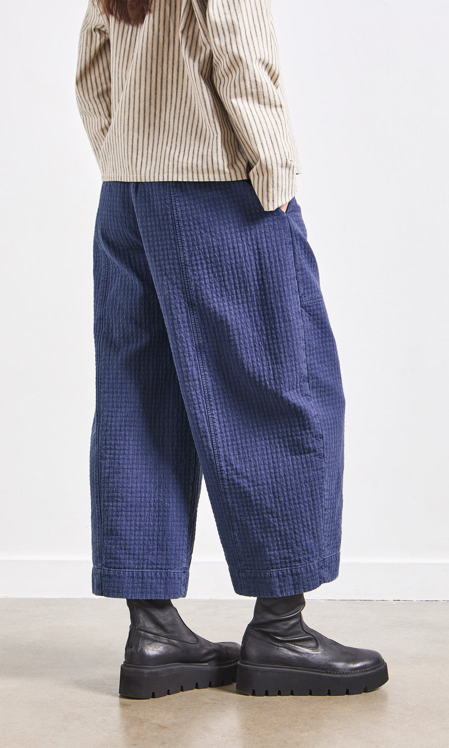 Plümo- Wearing blue checked Calow Pants, a beige striped shirt, and black chunky boots, standing indoors.