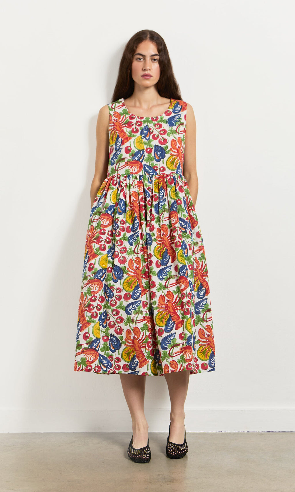Plümo- Woman in the Corallo Dress—colorful, hand-printed floral cotton voile with pockets and shell buttons—by a white wall.