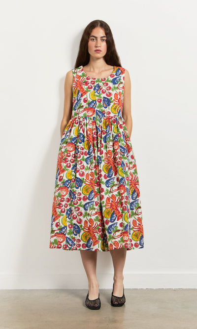 Plümo- Woman in the Corallo Dress—colorful, hand-printed floral cotton voile with pockets and shell buttons—by a white wall.