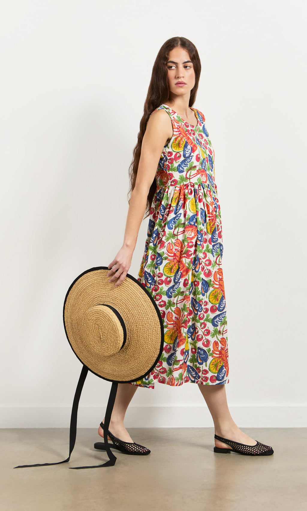 Plümo- Woman in the Corallo Dress holds a straw hat, standing against a plain white wall.