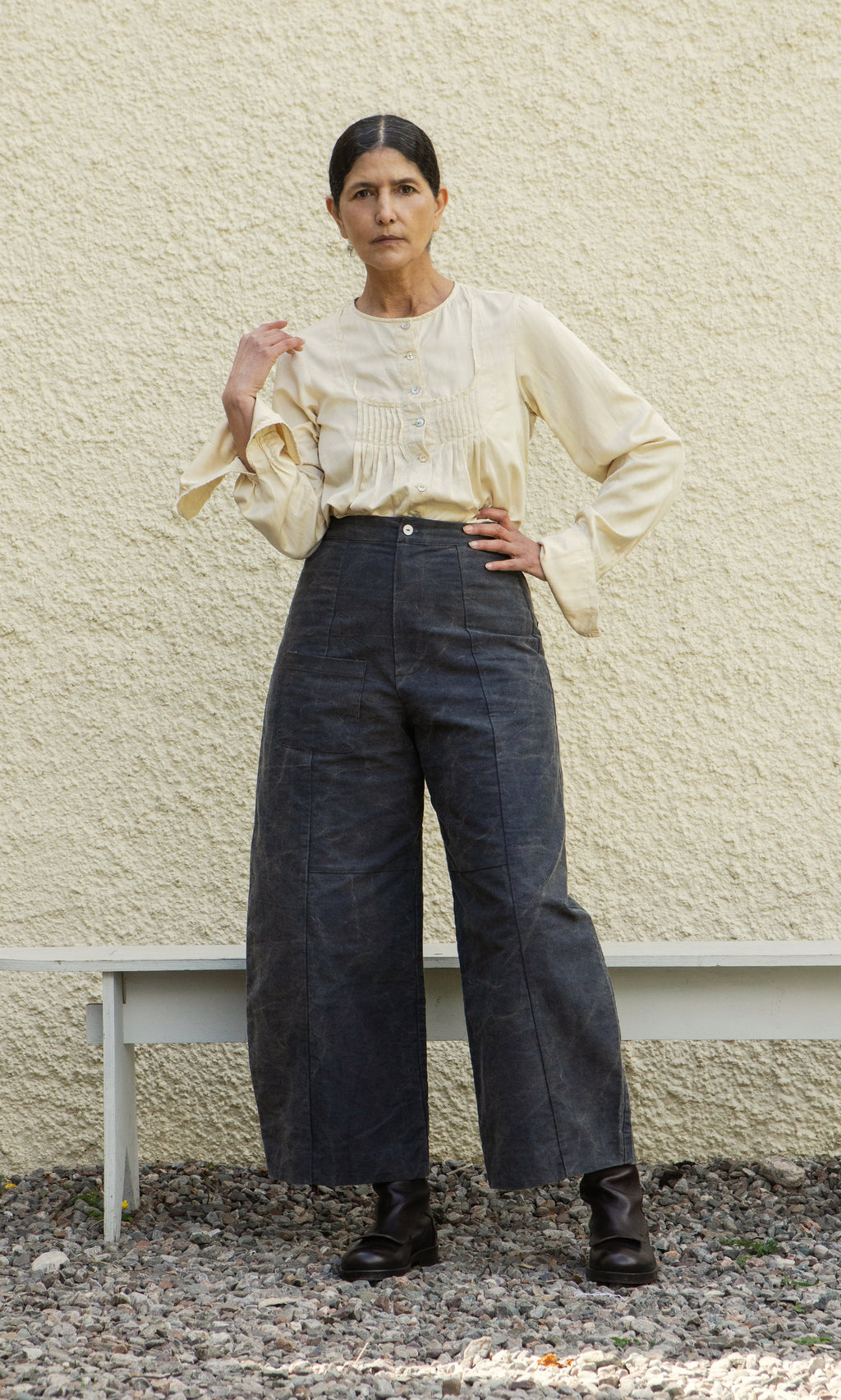 Plümo  Woman stands before a textured wall, wearing Corfe Barrel Pants, a cream blouse, and black boots.