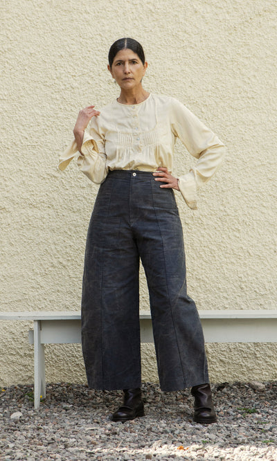 Plümo  Woman stands before a textured wall, wearing Corfe Barrel Pants, a cream blouse, and black boots.