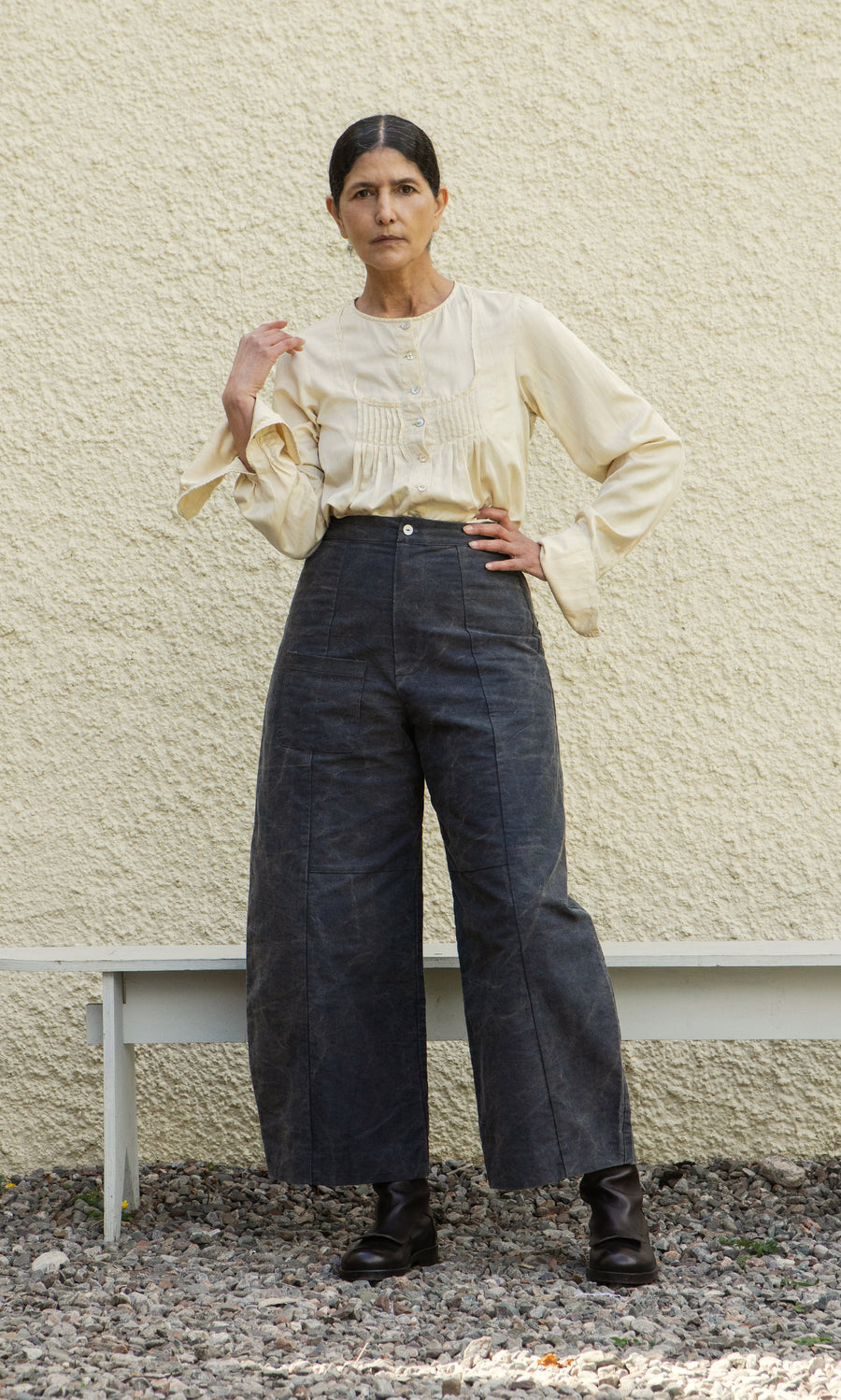 Plümo  Woman stands before a textured wall, wearing Corfe Barrel Pants, a cream blouse, and black boots.